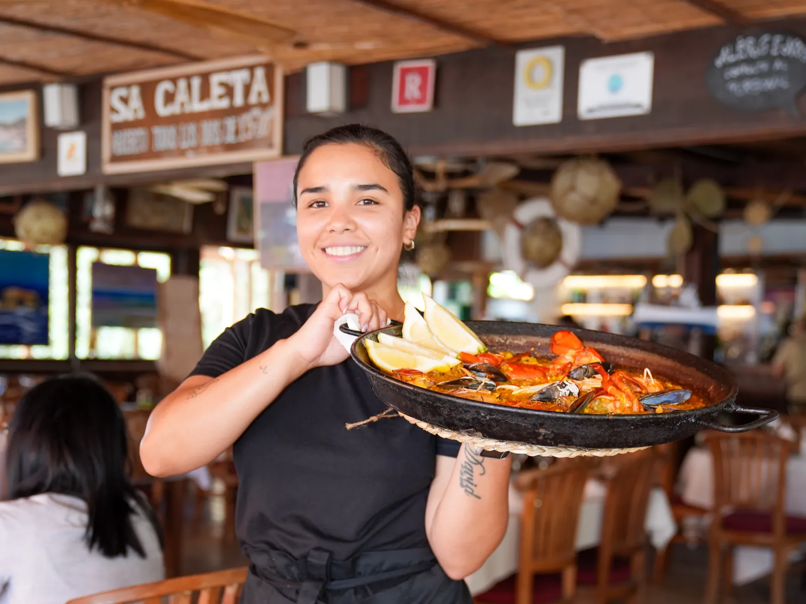 Camarera con una paella de marisco bajo el cartel de Sa Caleta